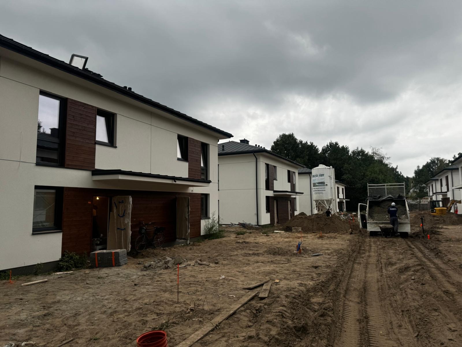 Nowoczesne domy z elewacją w kolorze złamanej bieli i elementami z ciemnego drewna. Na pierwszym planie plac budowy z pracującym robotnikiem i zaparkowaną ciężarówką.