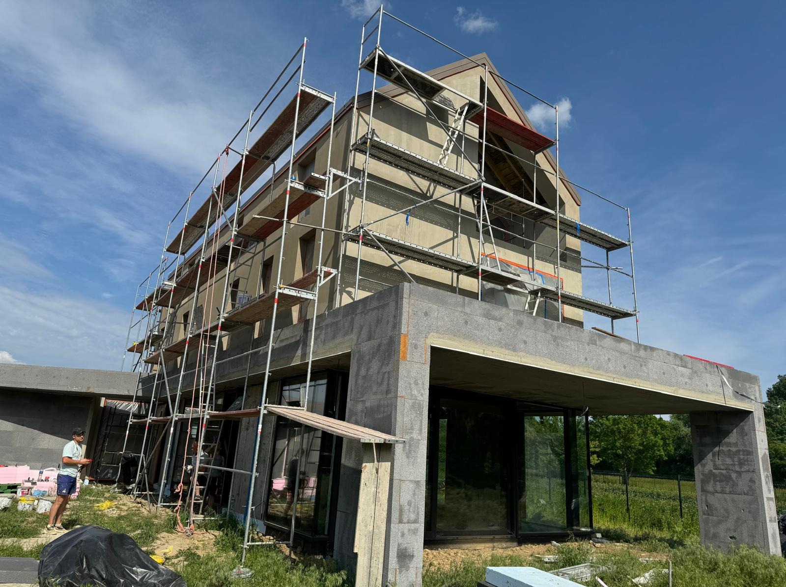 Dom w trakcie ocieplania, z widocznymi rusztowaniami i pracownikiem w tle. Elewacja pokryta siatką i klejem, przygotowana do dalszych prac wykończeniowych.