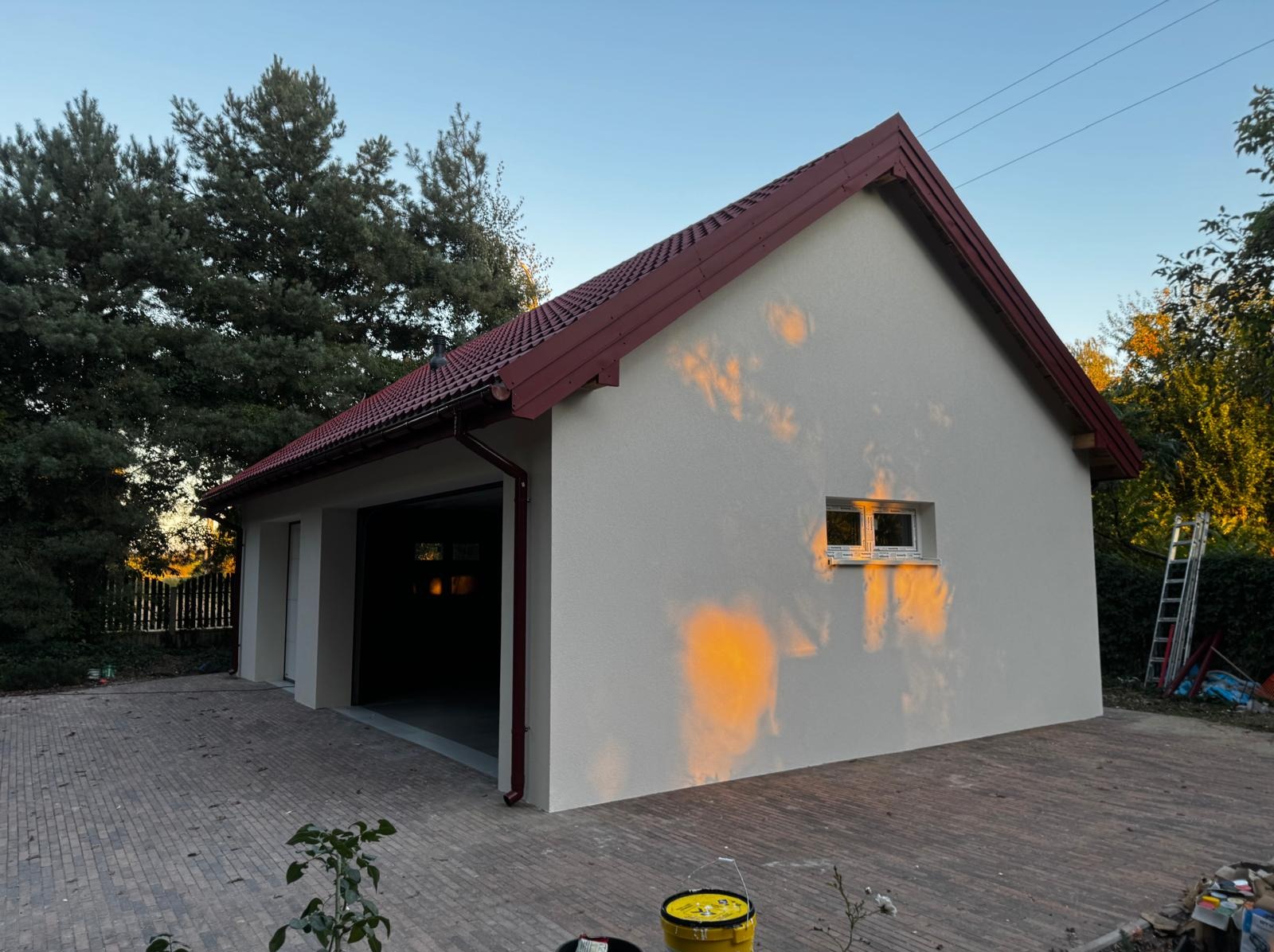 Świeżo otynkowany budynek gospodarczy z czerwoną dachówką, widoczne odbicie słońca na elewacji i wiadro z farbą. Kostka brukowa przed budynkiem.
