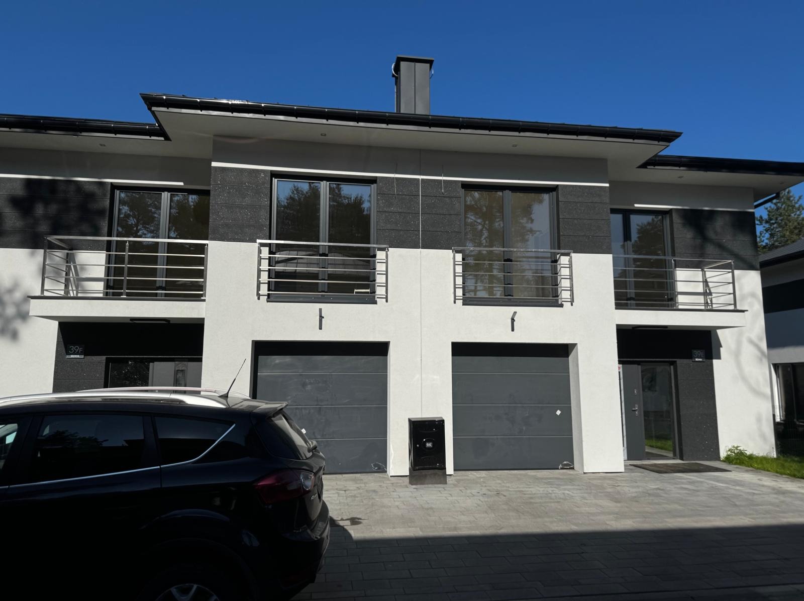 Nowoczesny dom bliźniak z grafitowo-białą elewacją, balkonami z metalowymi barierkami i dwoma garażami. Na pierwszym planie zaparkowany czarny samochód.