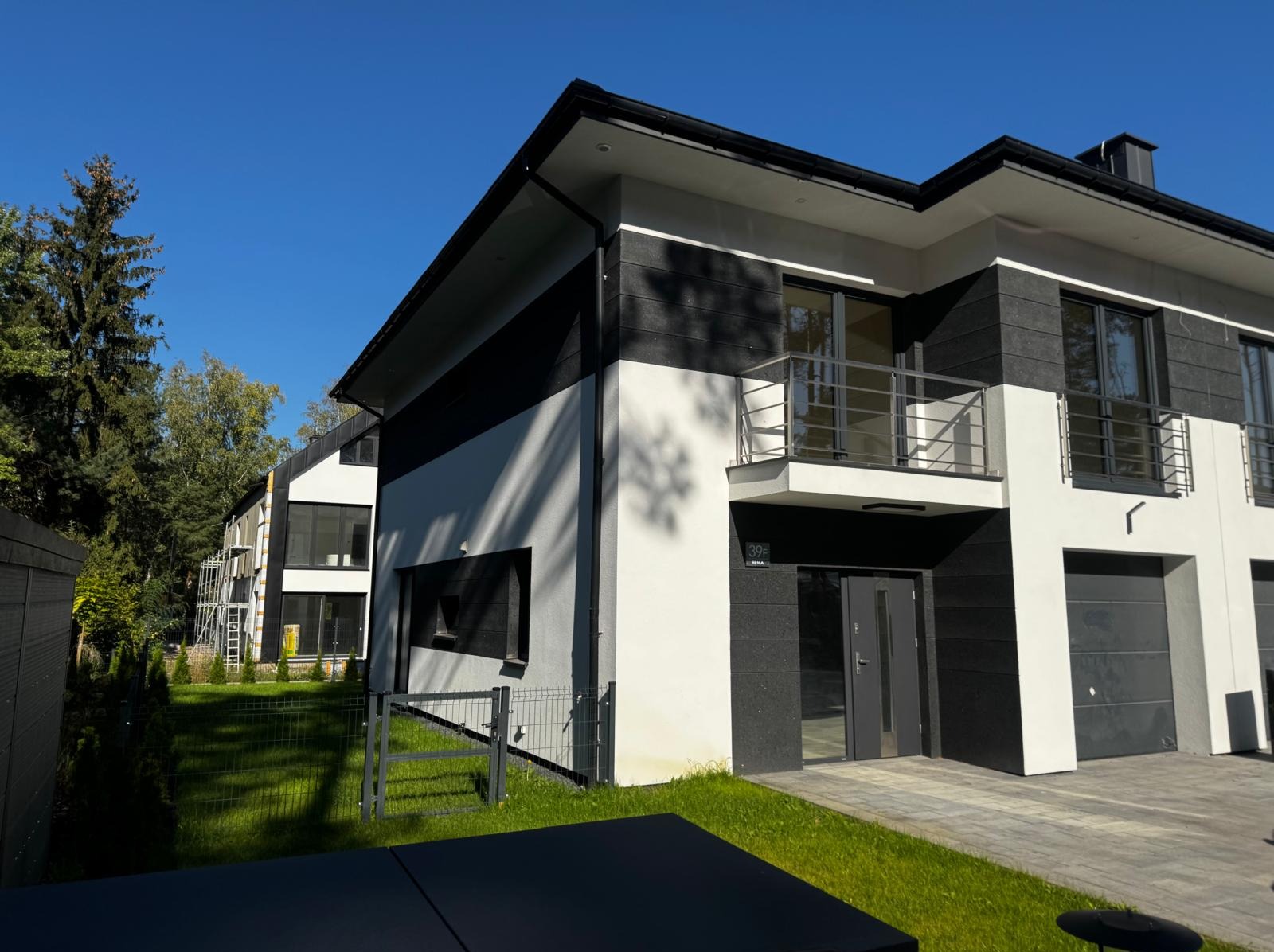 Nowoczesny dom z elewacją w odcieniach bieli i grafitu, z widocznymi detalami wykończenia, balkonem i fragmentem ogrodzenia. Na drugim planie dom w trakcie prac elewacyjnych.