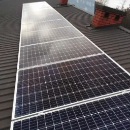 Rząd paneli słonecznych na ciemnym, blaszanym dachu domu jednorodzinnego, widoczne dwa kominy z cegły i fragment krajobrazu w tle.