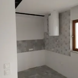 Wnętrze kuchni w trakcie remontu: szare płytki na podłodze, częściowo położone płytki ścienne z geometrycznym wzorem, biały okrągły okap i drewniane okno.