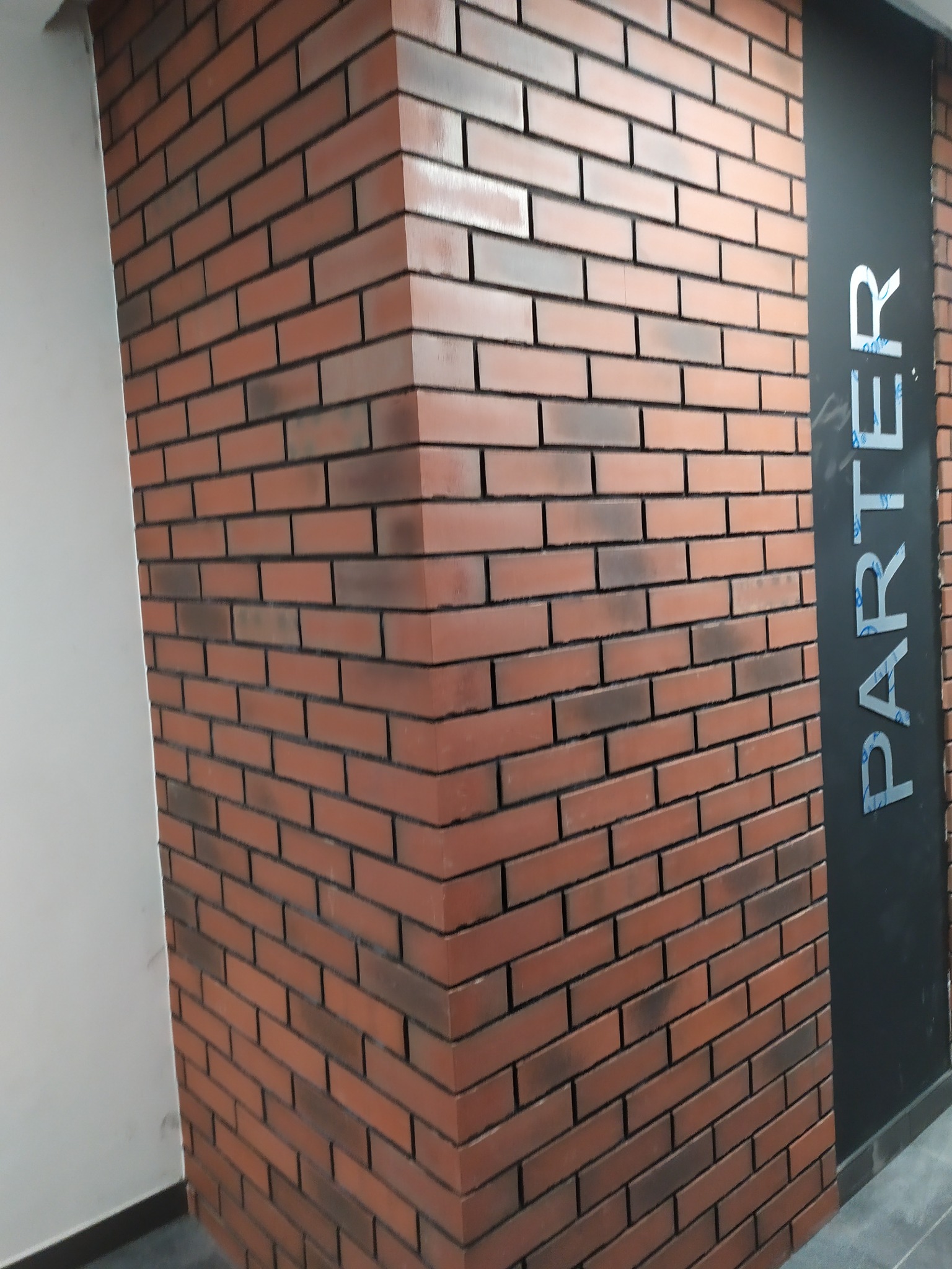 Wykończona ściana z cegieł klinkierowych w odcieniach czerwieni i brązu, narożnik ściany z widocznymi spoinami, obok czarna ściana z napisem 'PARTER' w srebrnych literach.