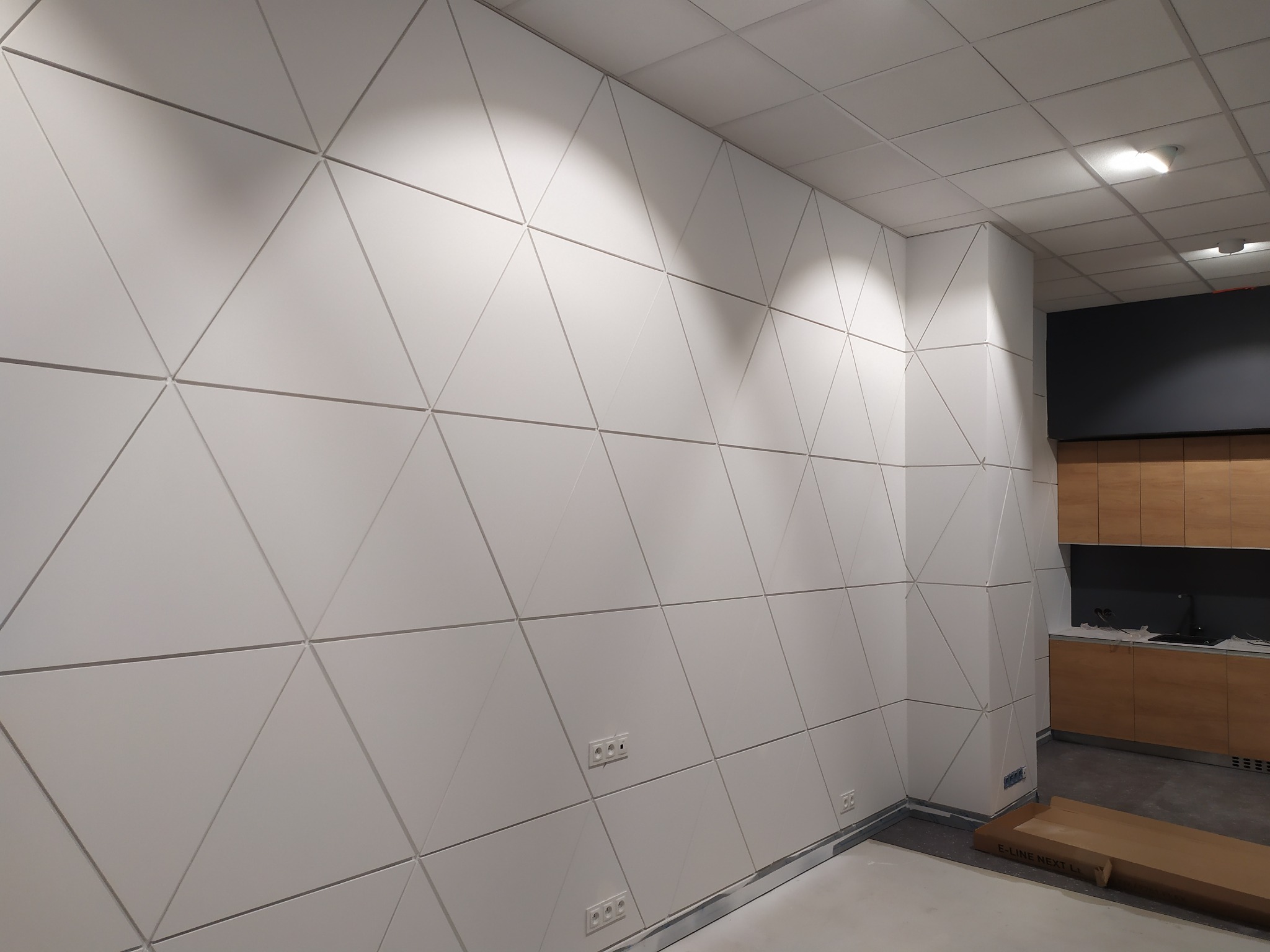Biała ściana z trójkątnymi panelami 3D, widoczne gniazdka elektryczne i fragment zabudowy kuchennej z drewnianymi szafkami