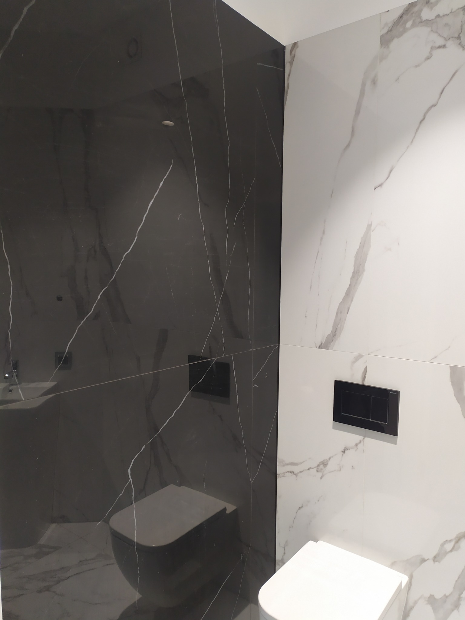 Nowoczesna łazienka z czarno-białymi marmurkowymi płytkami, wisząca toaleta z czarnym przyciskiem spłukującym, minimalistyczny design.