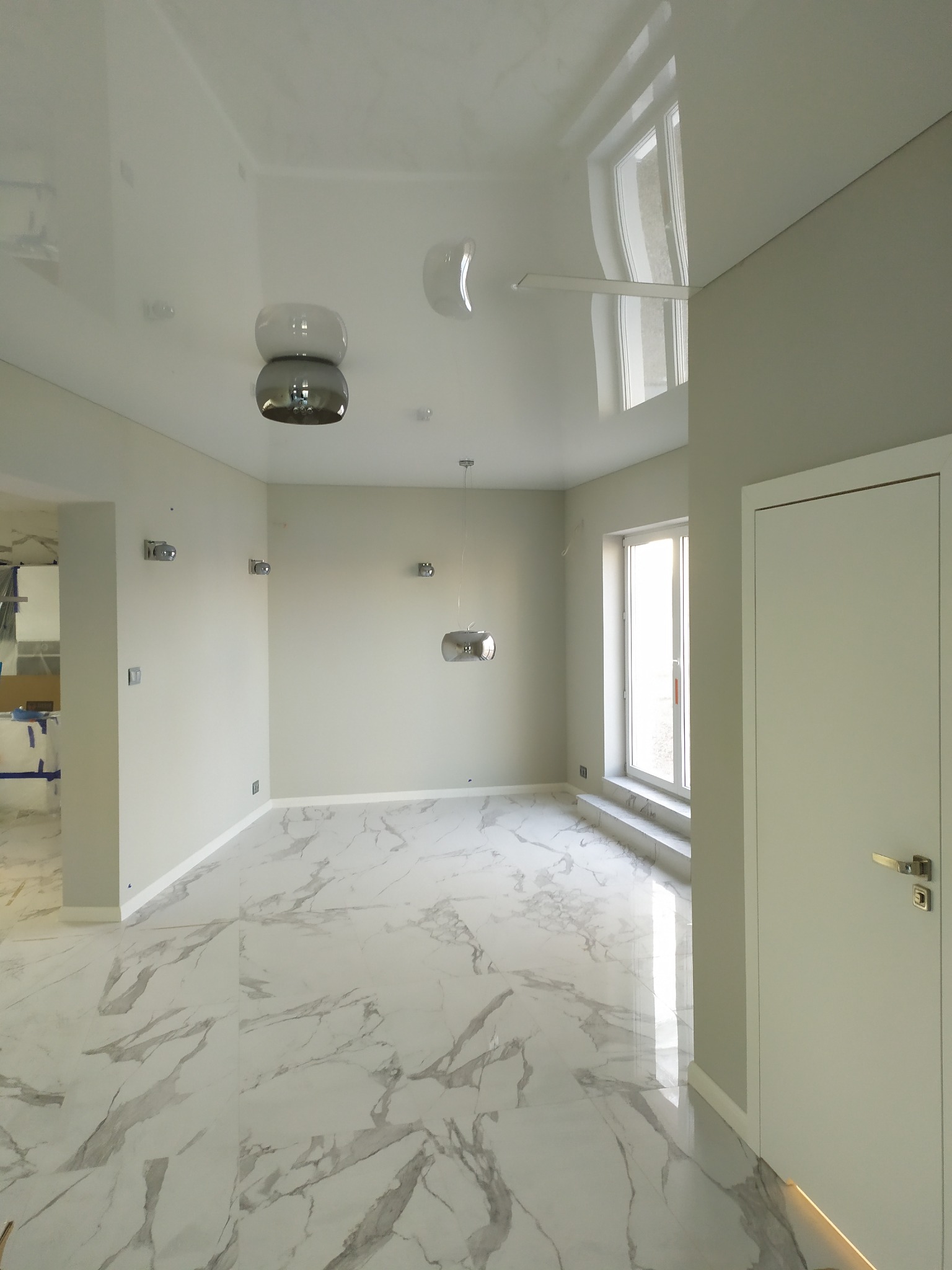 Jasne, przestronne wnętrze po remoncie: biała marmurowa podłoga z szarymi żyłami, błyszczący sufit z nowoczesnym oświetleniem, szare ściany, białe drzwi i okno z widokiem na zewnątrz.