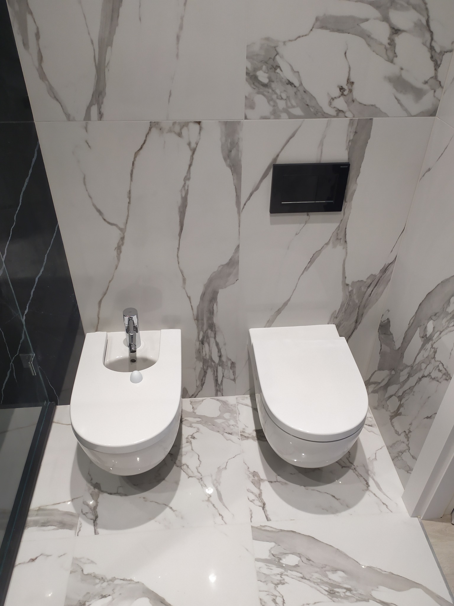 Nowoczesna łazienka w Warszawie z białymi płytkami imitującymi marmur, wisząca toaleta i bidet z chromowaną armaturą.