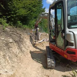 Mini koparka Takeushi TB225 z operatorem w trakcie pracy na zboczu, przygotowanie terenu pod instalacje, widoczne wykopane rury.