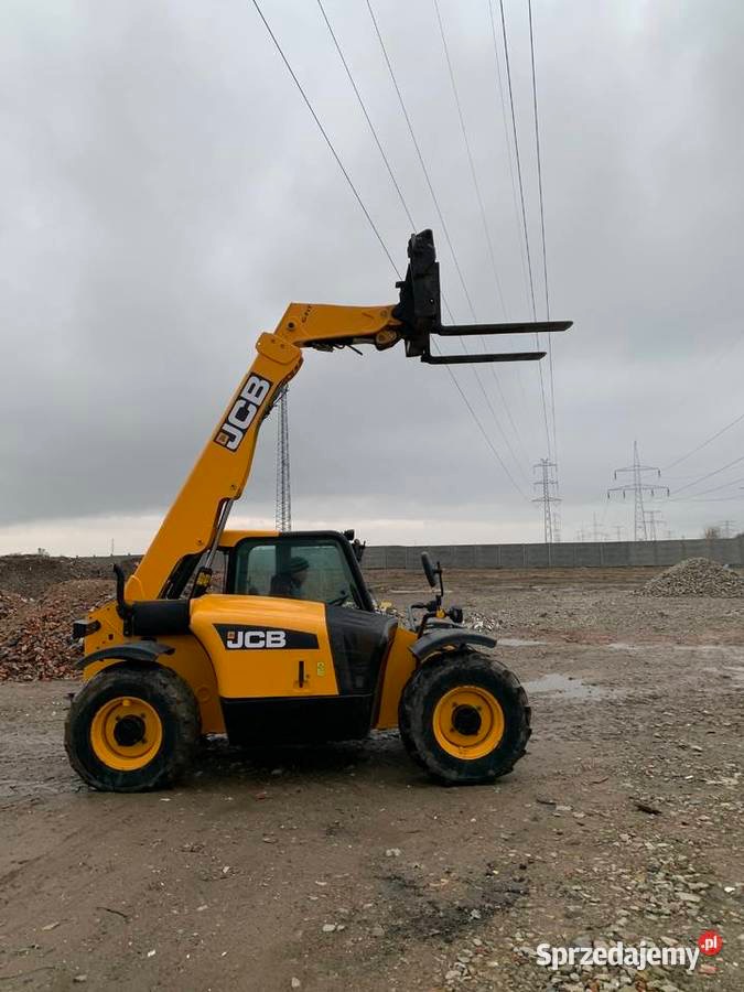 Żółto-czarny wózek teleskopowy JCB z podniesionym ramieniem i widłami na nierównym terenie budowy, widoczne linie wysokiego napięcia w tle.