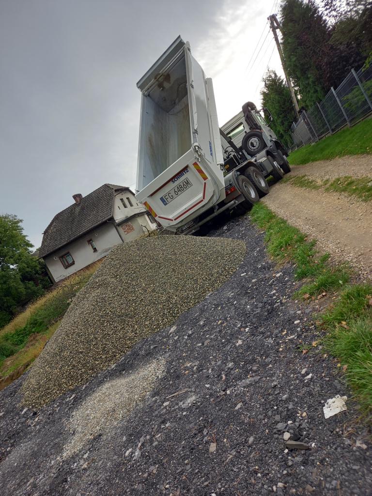 Wyładowany materiał sypki z wywrotki Schmitz na nierównym terenie, obok starego domu z ceglanym dachem, widok z dołu na pochmurne niebo.