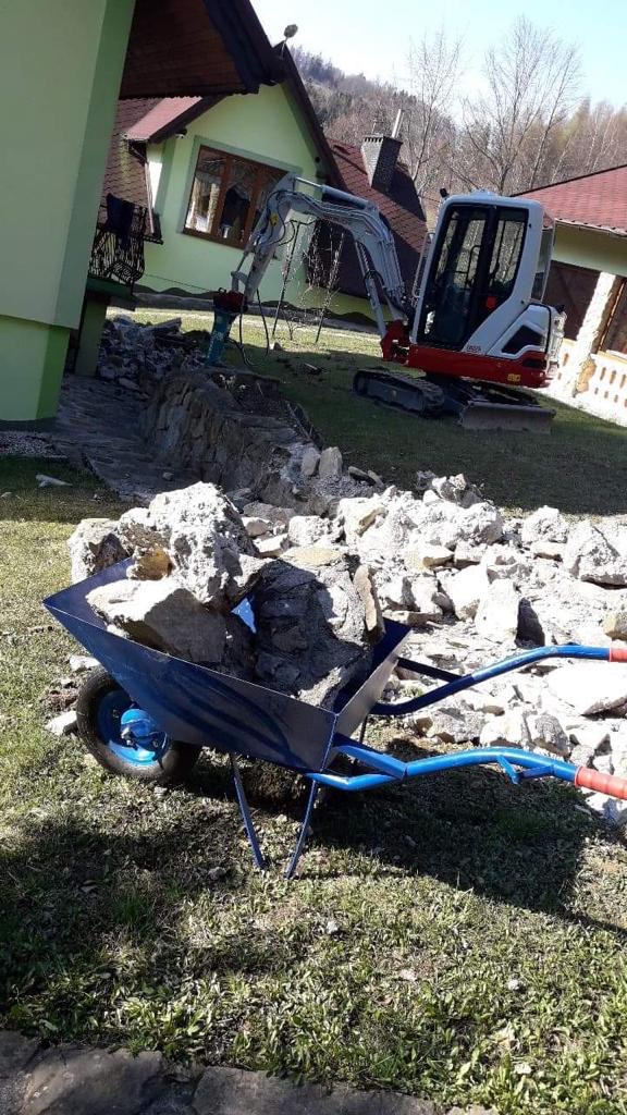 Niebieska taczka wypełniona gruzem skalnym stoi na trawie, w tle mała koparka przy budynku w trakcie prac ziemnych.