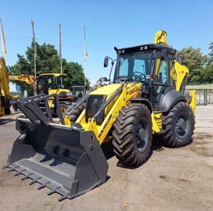 Żółta koparko-ładowarka New Holland B115B z przodu, z widoczną łyżką i oponami, na placu z innymi maszynami w tle, w słoneczny dzień.
