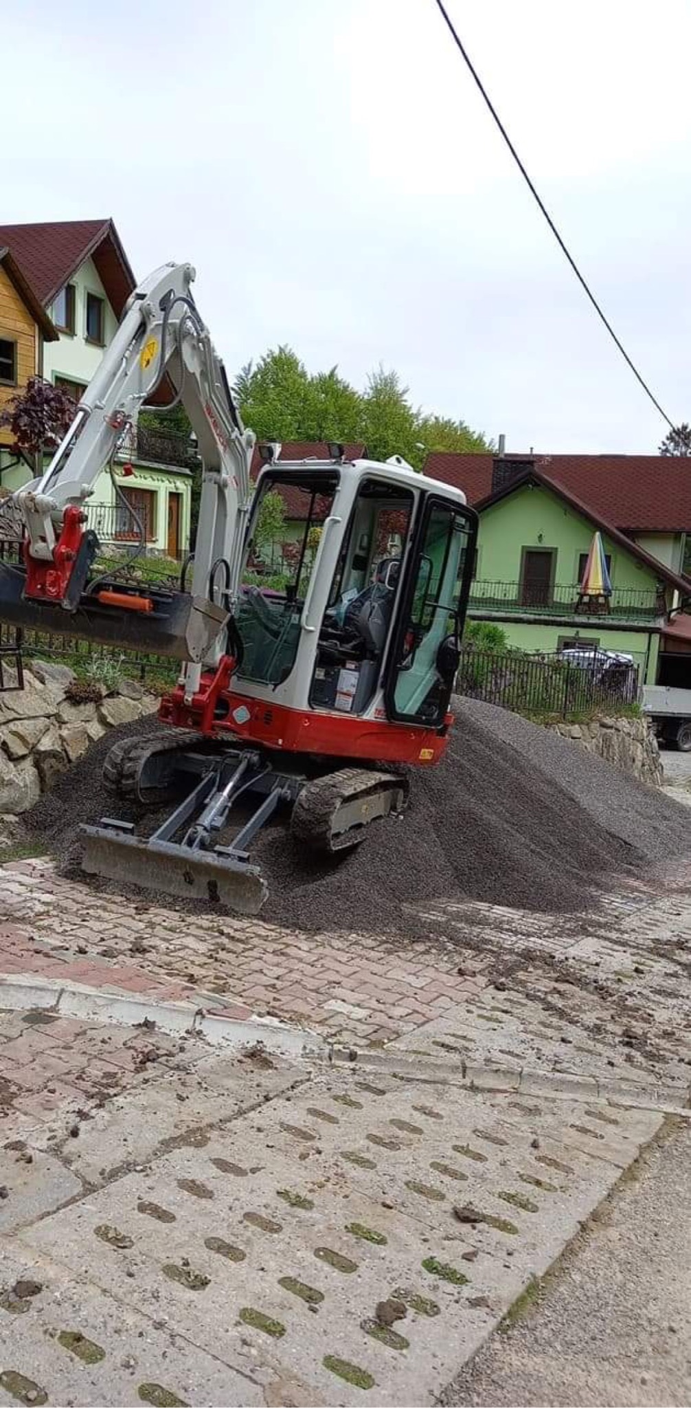 Mała koparka gąsienicowa z łyżką na tle hałdy żwiru, widoczne budynki mieszkalne w tle.