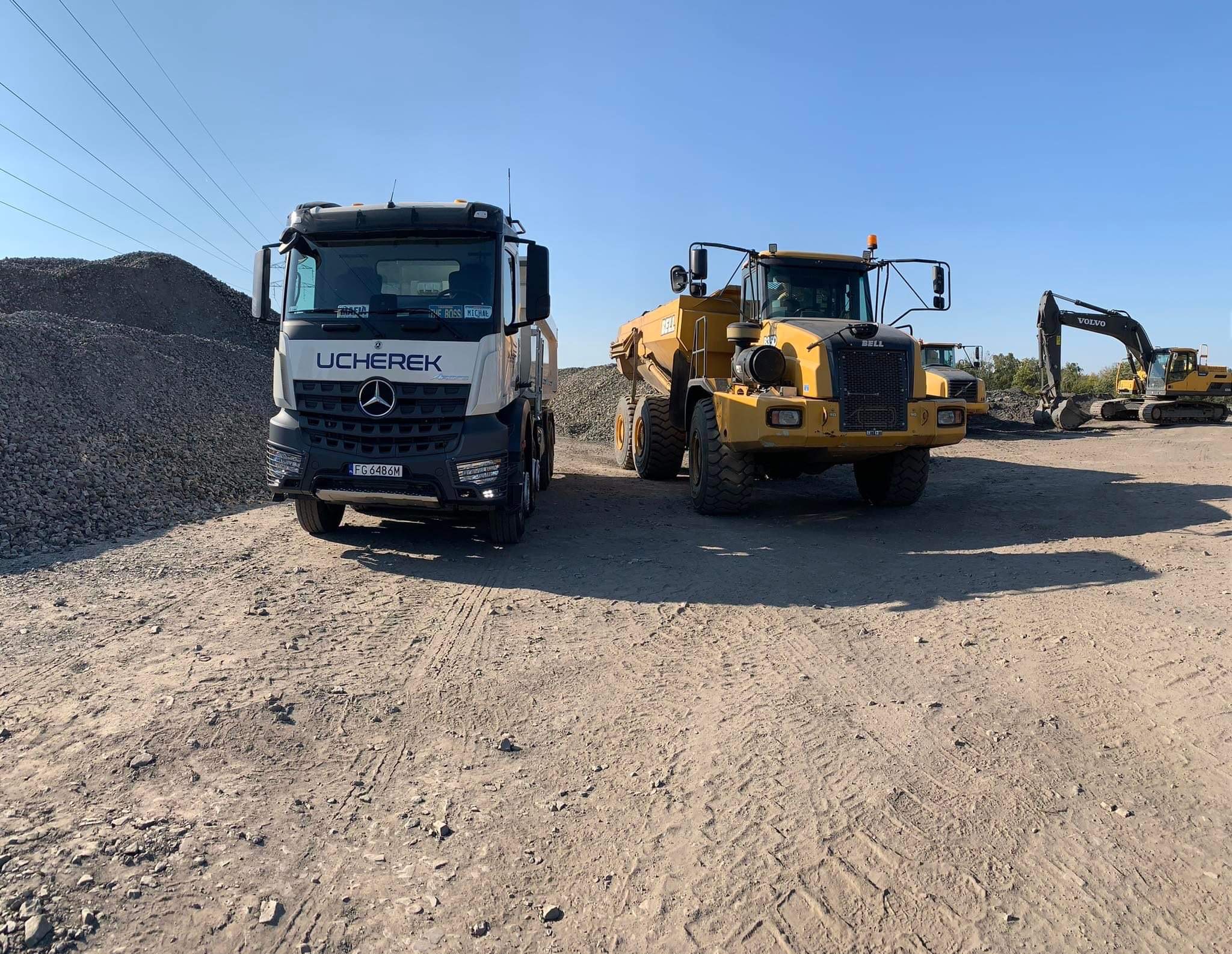 Ciężarówka Mercedes UCHEREK i żółty wozidło Bell B35D na placu budowy, w tle hałda gruzu i koparka Volvo