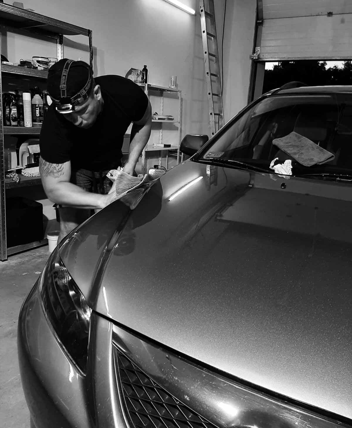 Mężczyzna z lampką czołową poleruje maskę szarego samochodu w garażu, widoczne półki z preparatami i drabina.