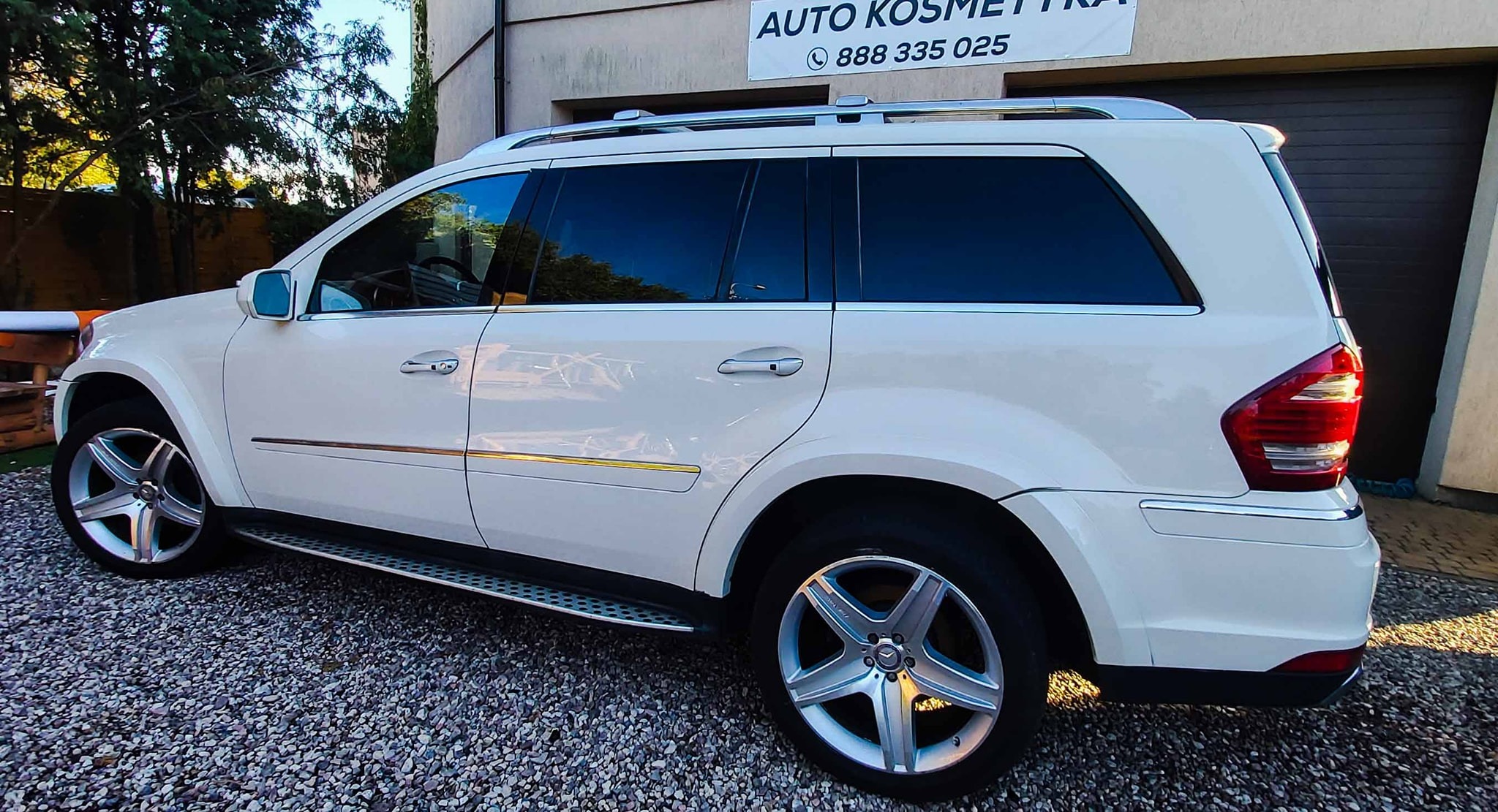 Biały samochód typu SUV marki Mercedes-Benz po czyszczeniu, zaparkowany na żwirowym podjeździe z widocznym logo firmy Auto Kosmetyka w tle.