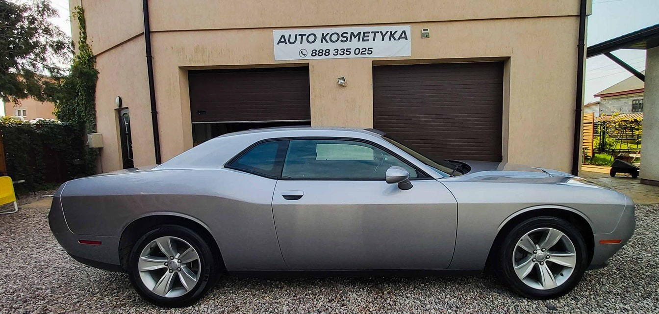 Srebrny Dodge Challenger po autokosmetyce, zaparkowany przed budynkiem z szyldem 'Auto Kosmetyka'.