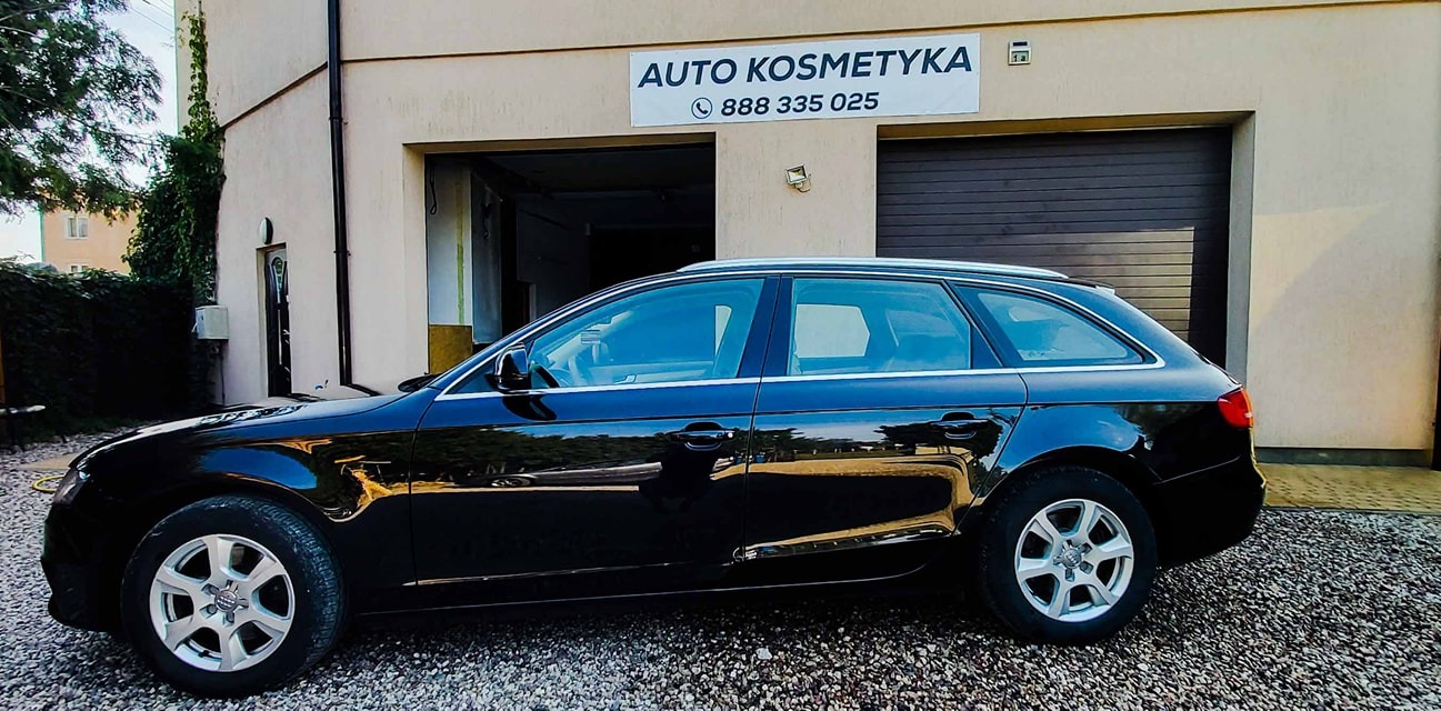 Czarny samochód kombi marki Audi zaparkowany przed budynkiem auto kosmetyki z widocznym szyldem reklamowym i numerem telefonu.