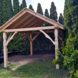 Surowa drewniana konstrukcja altany ogrodowej z ozdobnymi kolumnami i niezadaszonym dachem, otoczona gęstymi tujami i trawnikiem.