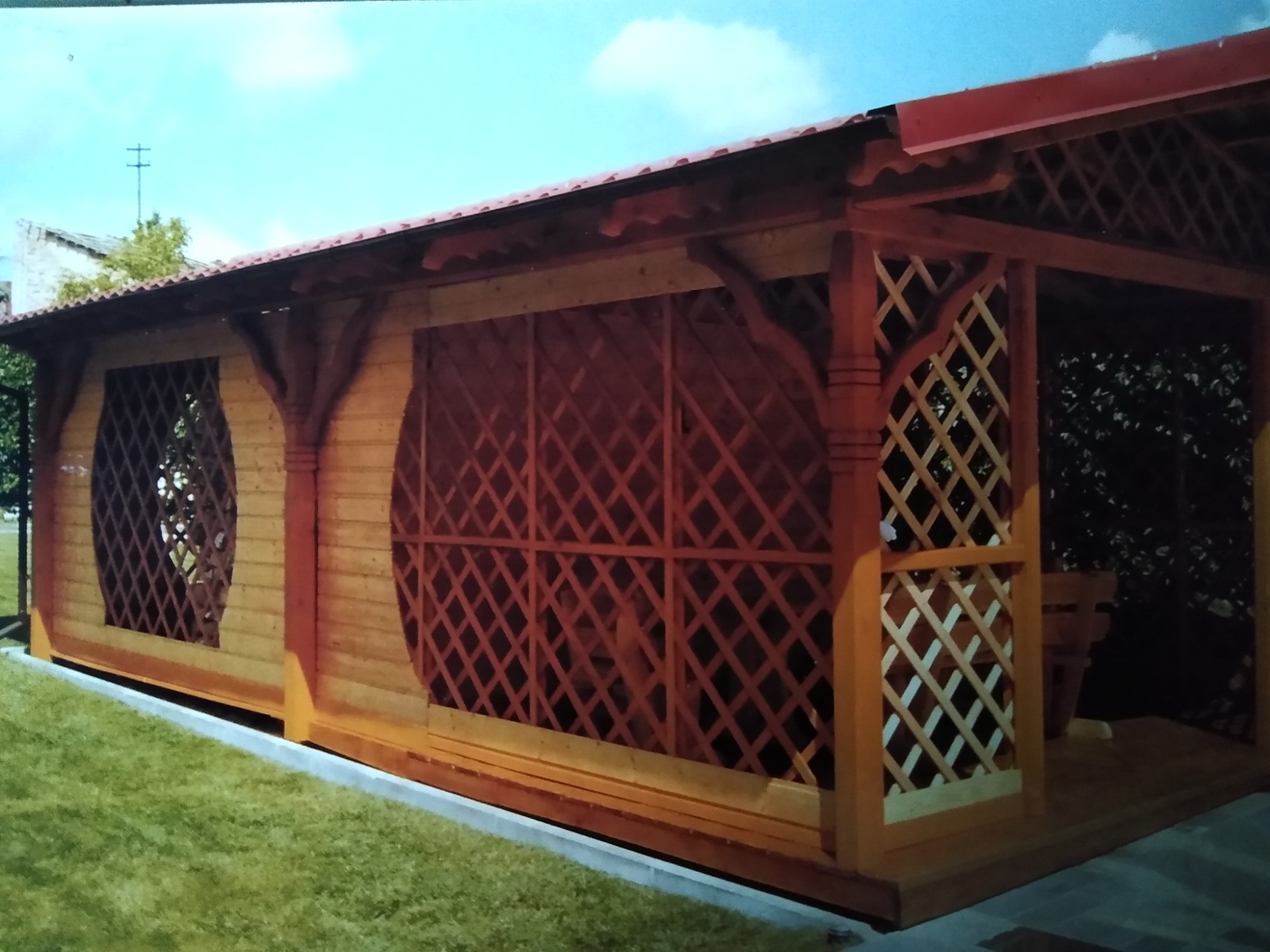 Drewniana konstrukcja pawilonu ogrodowego z kratownicowymi ścianami, czerwonym dachem i dekoracyjnymi kolumnami, otoczona trawnikiem.
