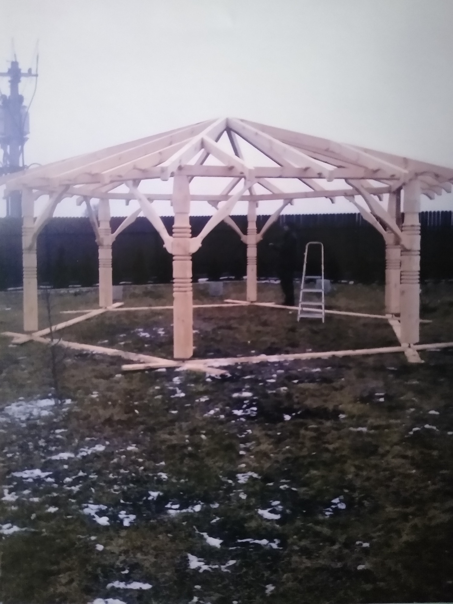 Konstrukcja drewnianej pergoli w trakcie budowy, widoczna drabina i częściowo pokryta śniegiem trawa.