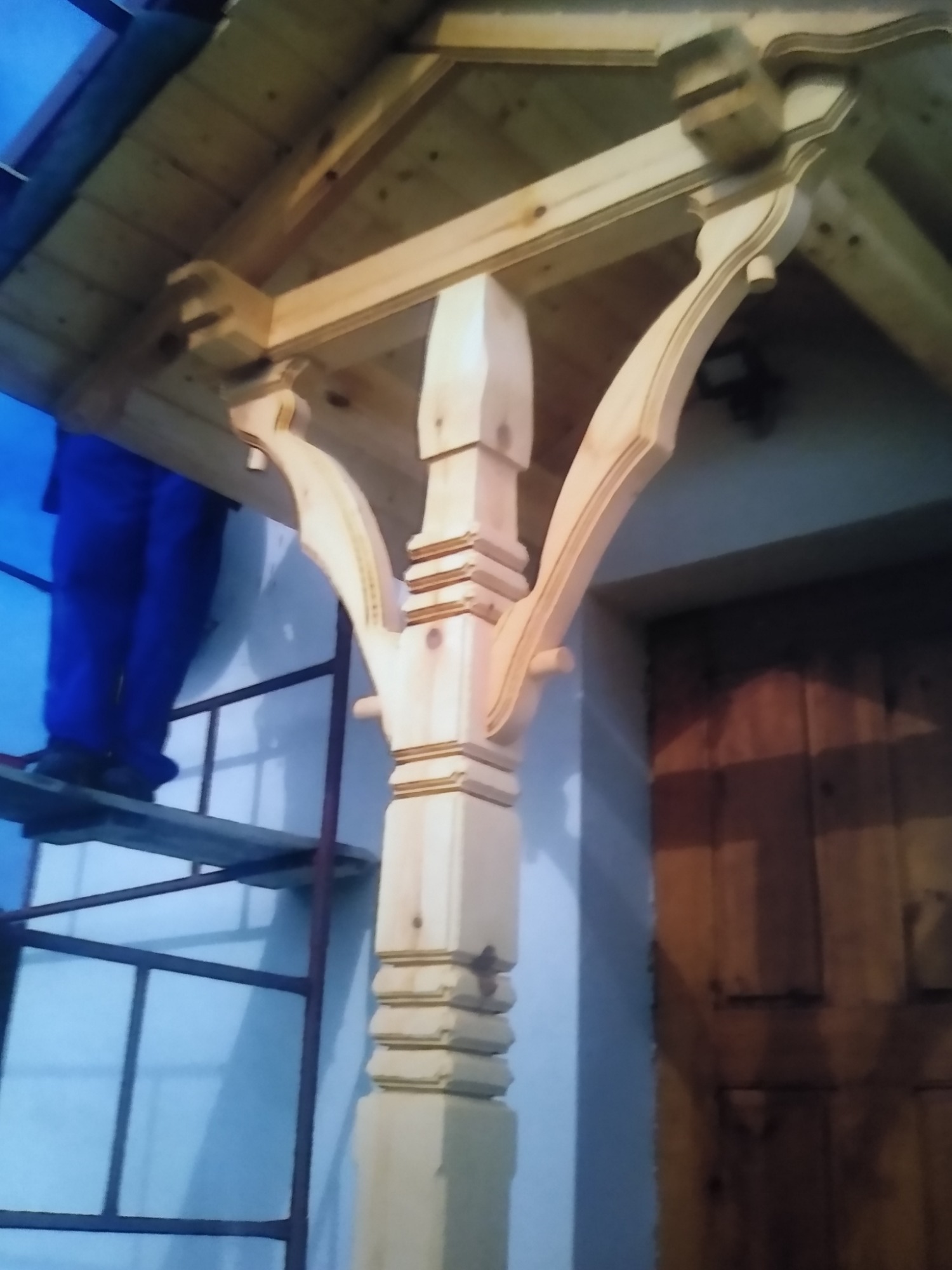 Drewniana kolumna z dekoracyjnymi elementami podtrzymująca zadaszenie werandy, widoczna osoba na rusztowaniu w trakcie montażu.