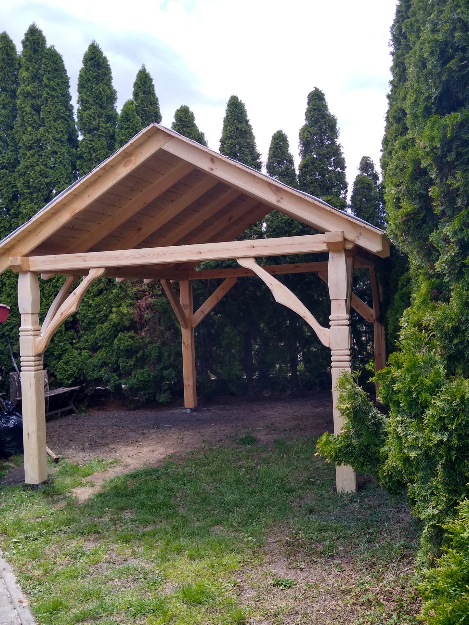 Surowa drewniana konstrukcja altany ogrodowej z ozdobnymi kolumnami i niezadaszonym dachem, otoczona gęstymi tujami i trawnikiem.