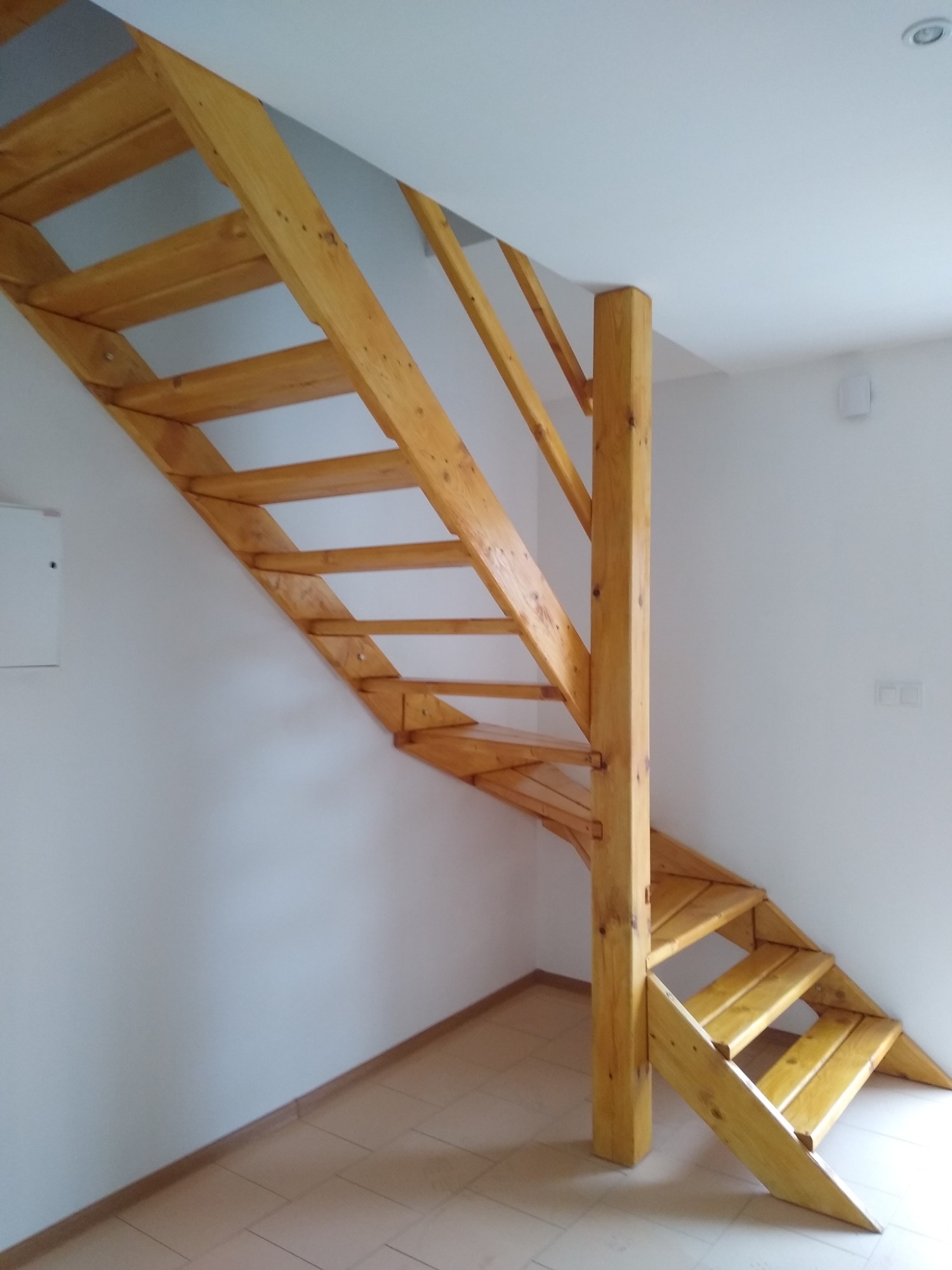 Drewniane schody ażurowe o jasnym odcieniu, z widoczną strukturą drewna, umieszczone w pomieszczeniu z białymi ścianami i jasną podłogą z płytek.