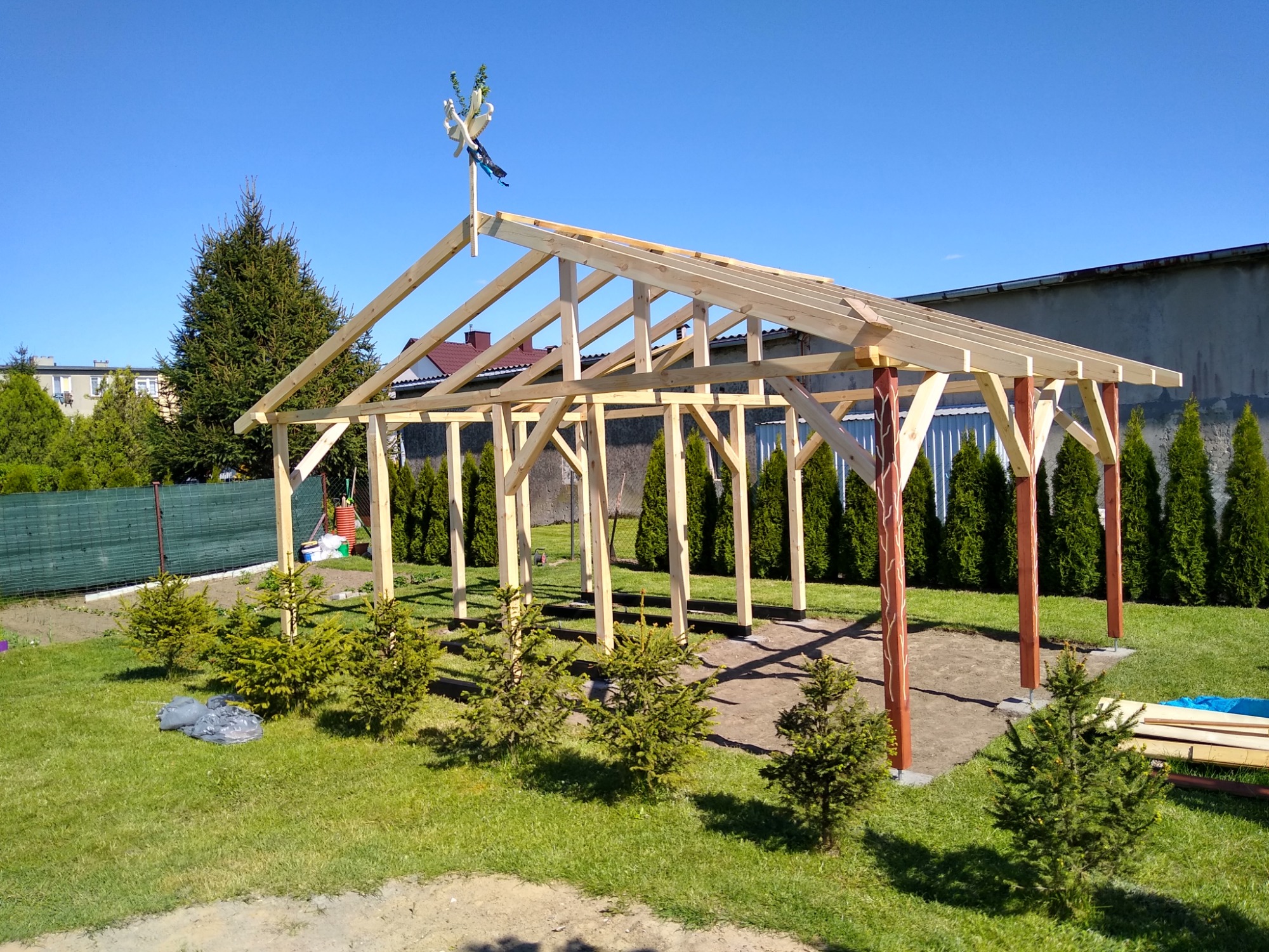 Konstrukcja drewnianej pergoli w trakcie budowy, z dekoracyjnym wieńcem na szczycie, widoczna na tle zielonego trawnika i wysokich tuj.