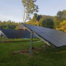 Dwa rzędy paneli słonecznych na metalowych stelażach, ustawione na trawniku przed budynkiem z blachy falistej; w tle zalesione wzgórza i niebo o zachodzie słońca, a po lewej stronie zaparkowany...