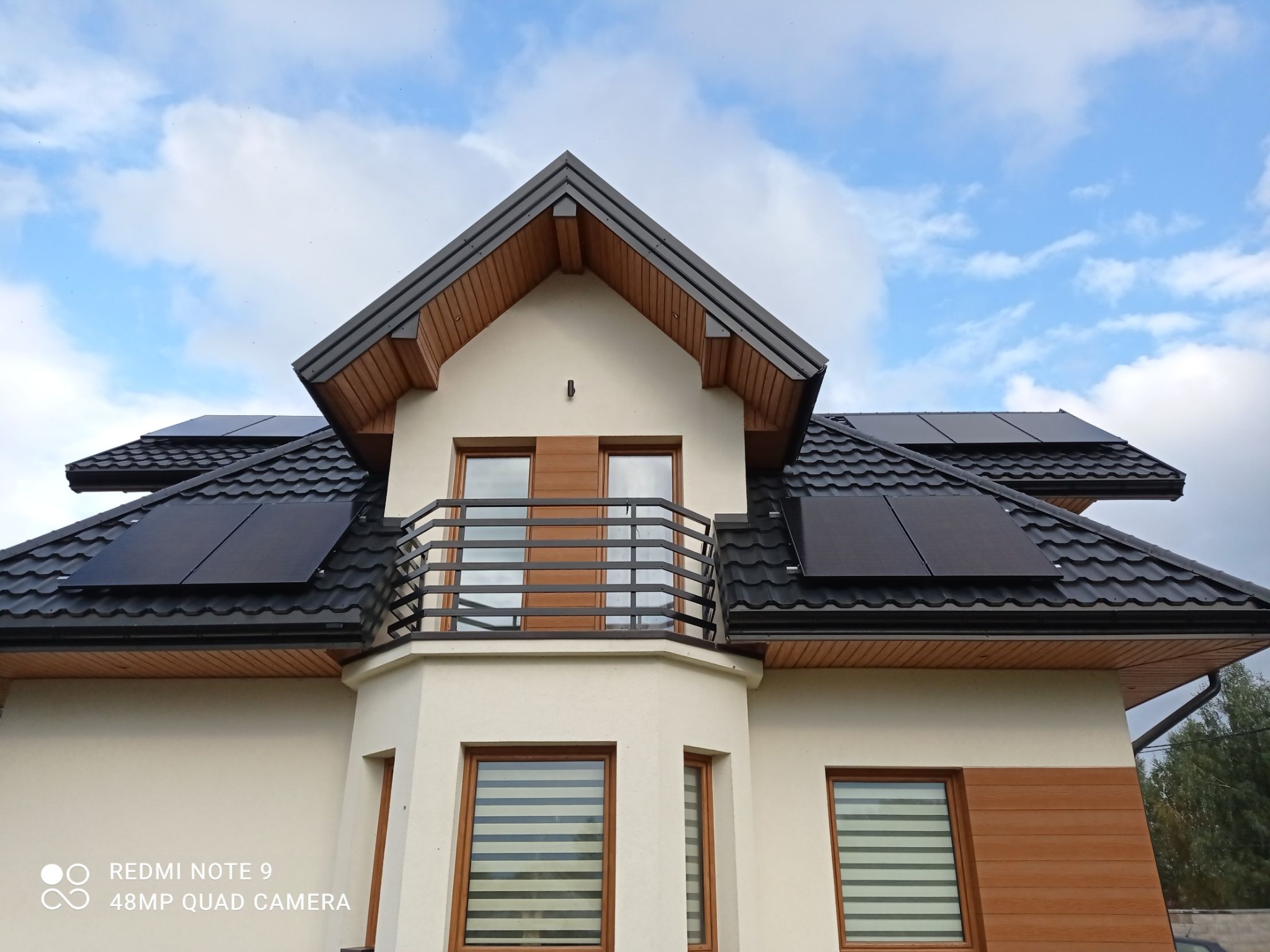 Panele słoneczne na dachu domu z balkonem, widok z dołu, błękitne niebo z chmurami w tle.