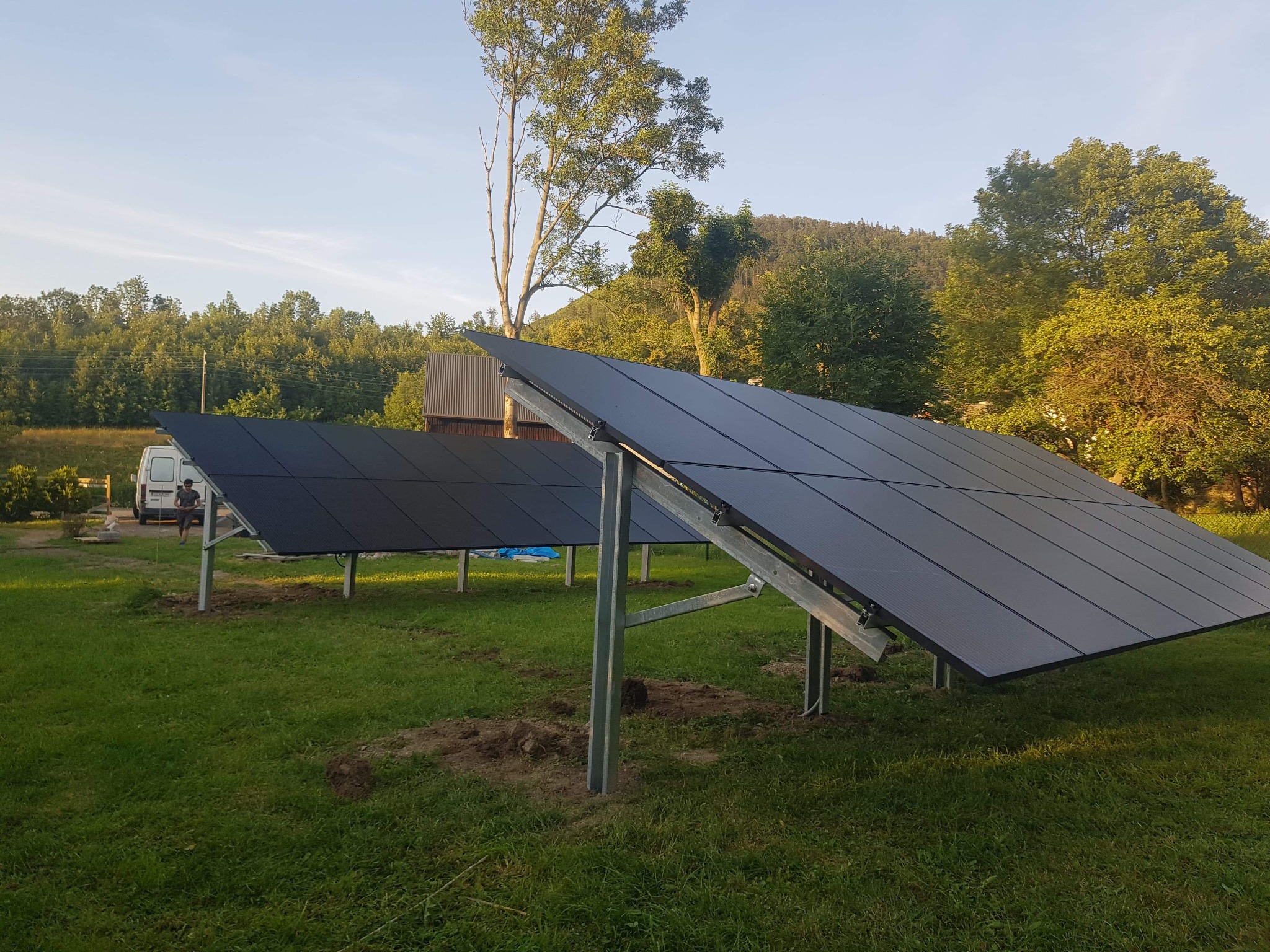 Dwa rzędy paneli słonecznych na metalowych stelażach, ustawione na trawniku przed budynkiem z blachy falistej; w tle zalesione wzgórza i niebo o zachodzie słońca, a po lewej stronie zaparkowany...