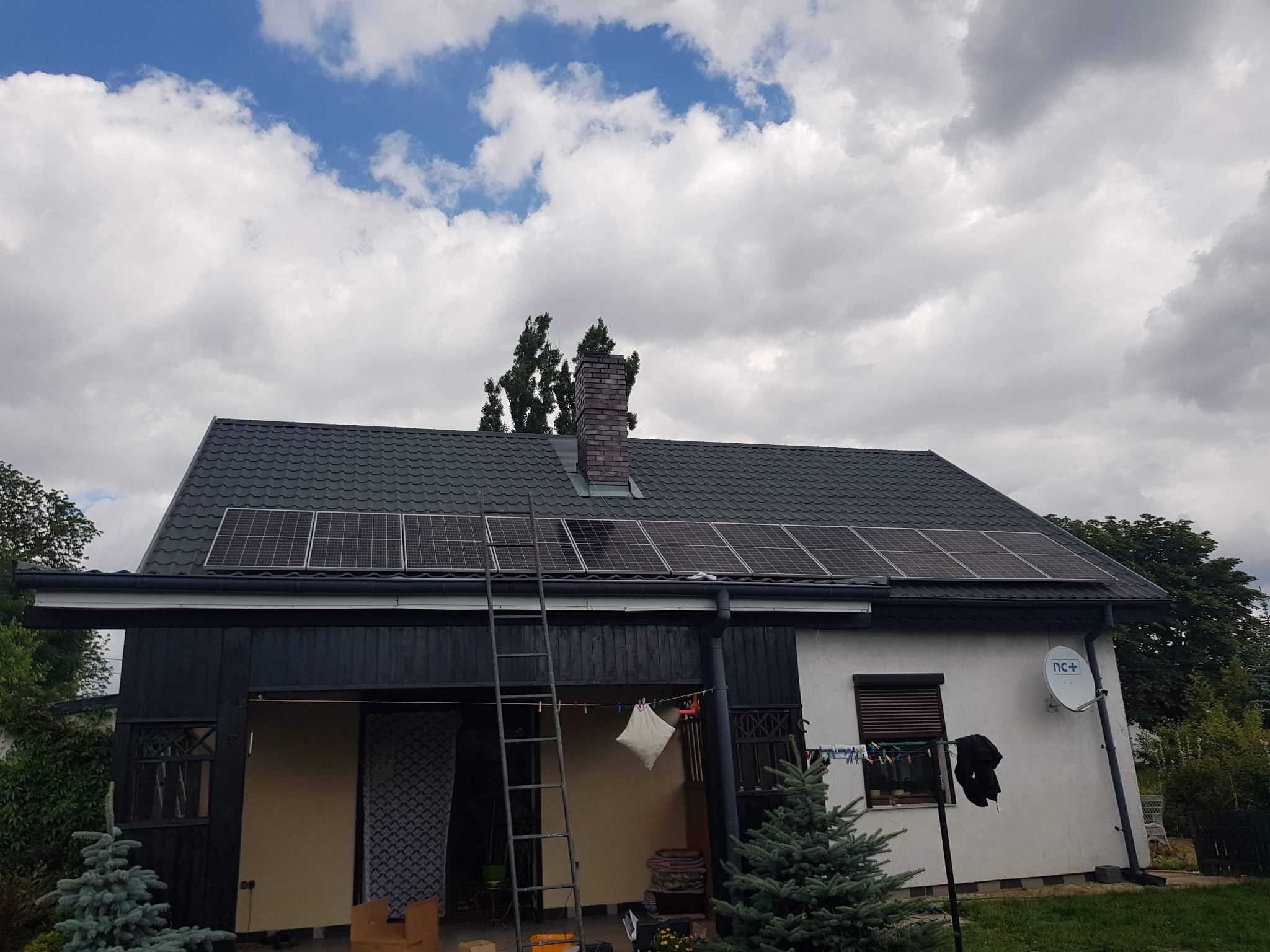 Dom jednorodzinny z zainstalowanymi panelami słonecznymi na dachu, drabina oparta o ścianę, widoczny komin z cegły i antena satelitarna na elewacji.