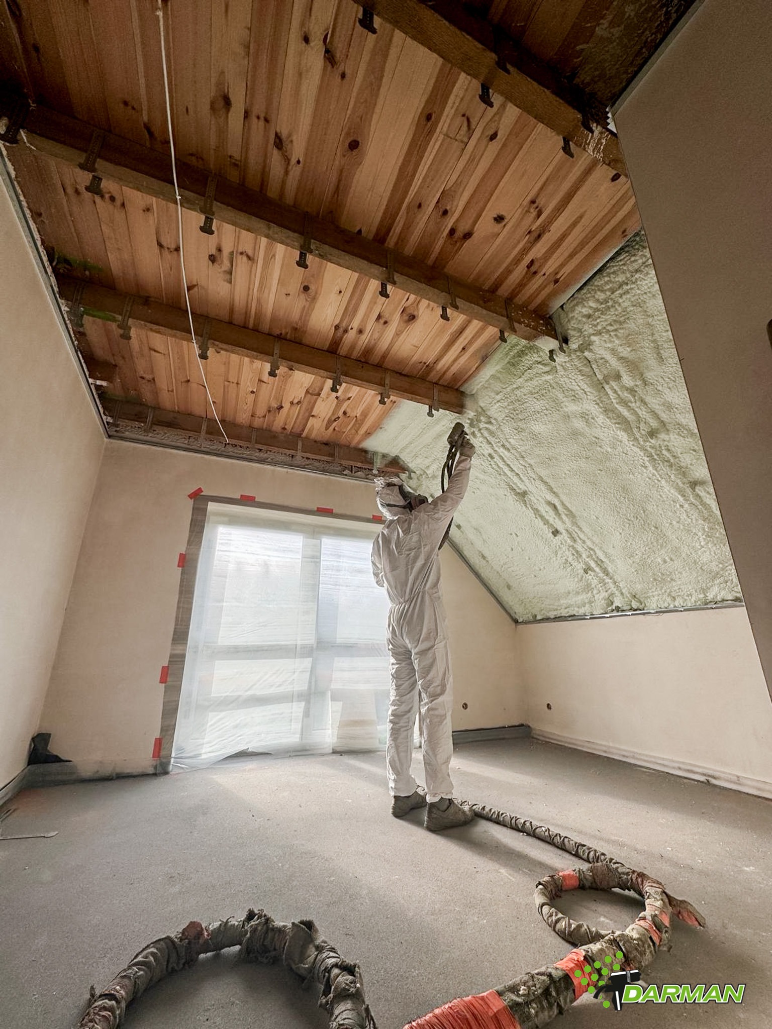 Pracownik w kombinezonie ochronnym natryskuje pianę izolacyjną na drewniany strop poddasza, widoczny wąż doprowadzający materiał i okno zabezpieczone folią.