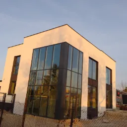 Nowoczesny dom z dużymi przeszkleniami, widoczny z zewnątrz przez metalowe ogrodzenie i elementy placu budowy, oświetlony popołudniowym słońcem.