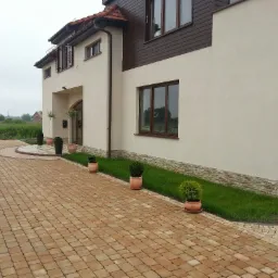 Beżowa kostka brukowa ułożona na podjeździe przed domem z jasną elewacją i brązowymi oknami, z dekoracyjnymi roślinami w donicach wzdłuż ścieżki i trawnika.