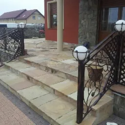 Schody zewnętrzne wyłożone piaskowcem z kutą balustradą i oświetleniem kulistym, prowadzące do wejścia budynku z kolumną i okładziną kamienną.