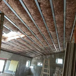 Wnętrze w trakcie ocieplania poddasza wełną mineralną, widoczne metalowe profile sufitowe, drabina i surowe ściany z oknami.