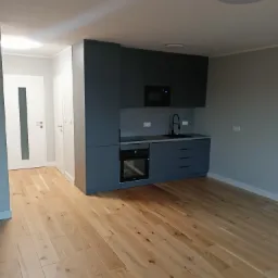 Nowoczesna, minimalistyczna kuchnia z szarymi szafkami, drewnianą podłogą i białymi drzwiami z przeszkleniem, widok z perspektywy wejścia.