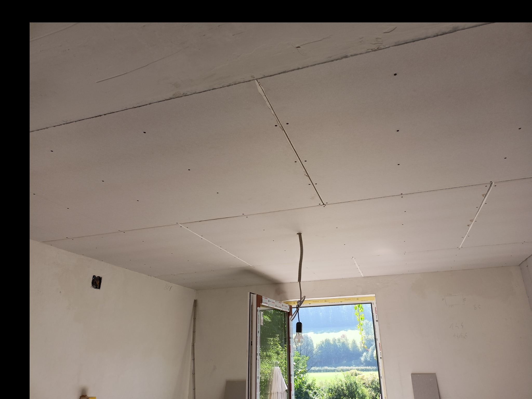 Sufit z płyt kartonowo-gipsowych w trakcie montażu, widoczne łączenia i wkręty, surowy kabel zwisający z otworu w suficie, okno z widokiem na zieleń w tle.