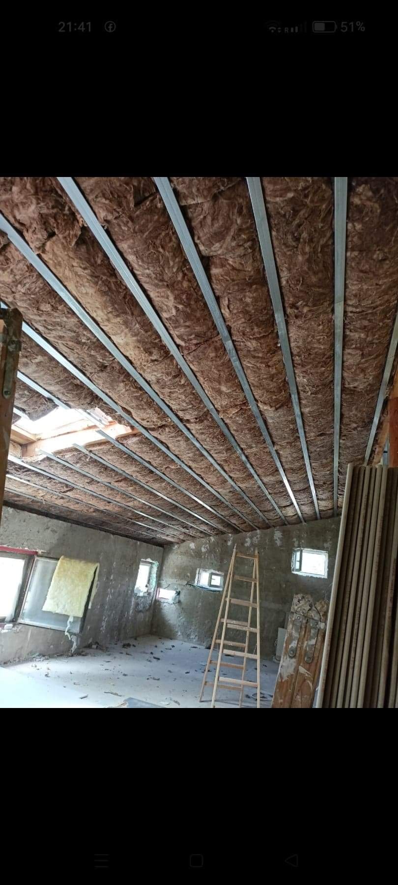 Wnętrze w trakcie ocieplania poddasza wełną mineralną, widoczne metalowe profile sufitowe, drabina i surowe ściany z oknami.