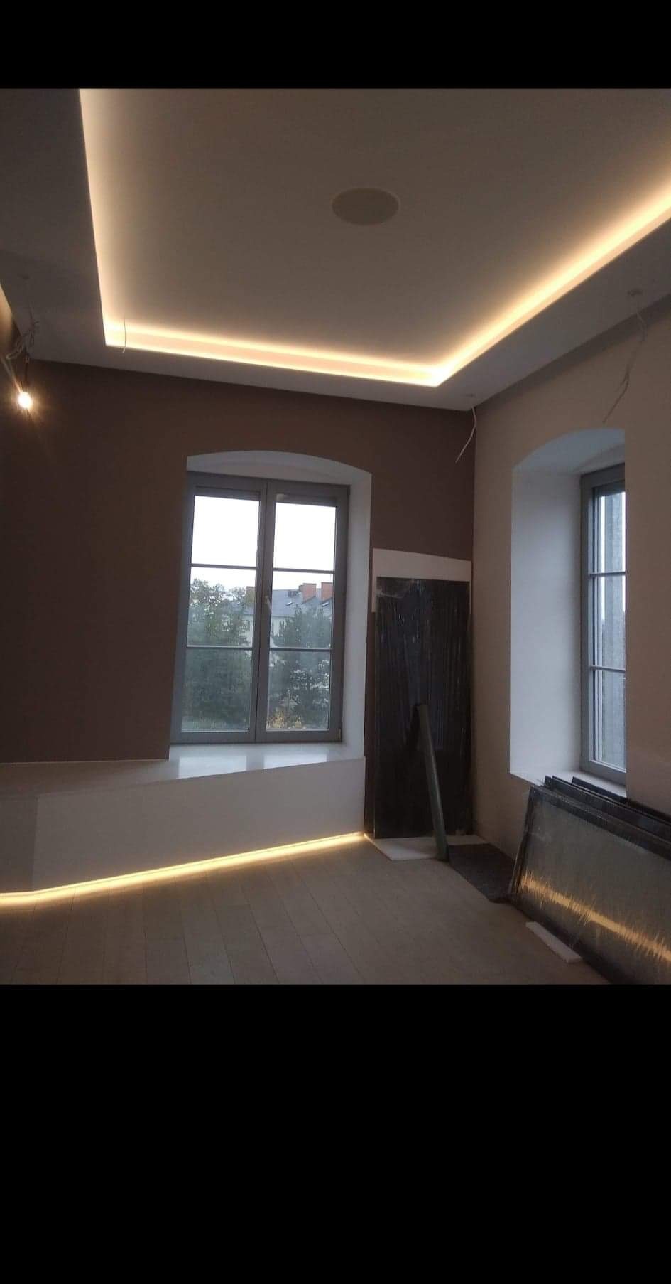 Wnętrze pokoju z dwoma oknami w szarych ramach, podświetleniem LED na suficie i wnęce okiennej, brązową ścianą i elementami w trakcie remontu, takimi jak folia ochronna na drzwiach i szyby oparte...