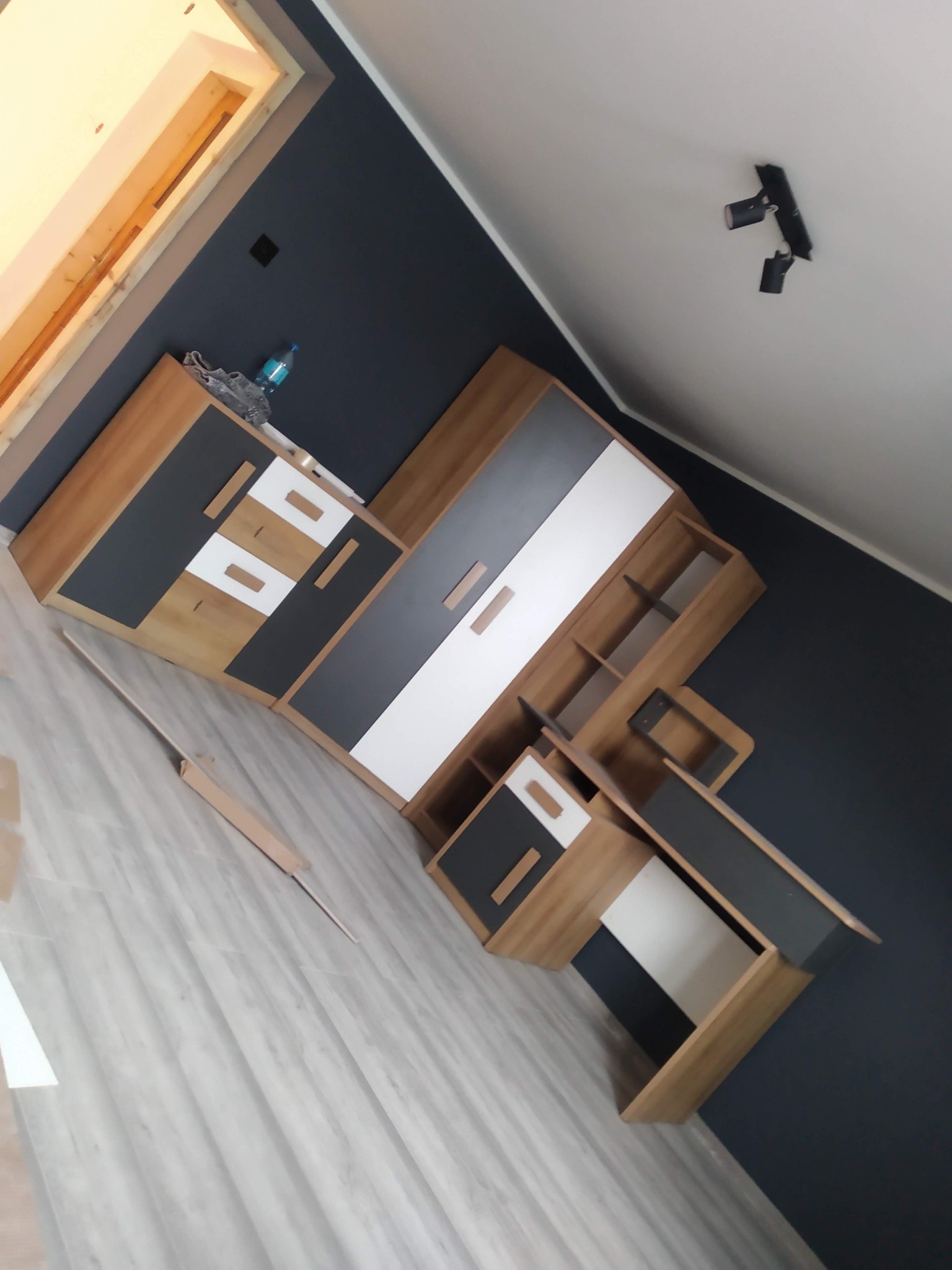 Zmontowane meble pokojowe w odcieniach drewna, szarości i bieli, z widocznym oświetleniem sufitowym i fragmentem listwy przypodłogowej, ustawione na panelach podłogowych.