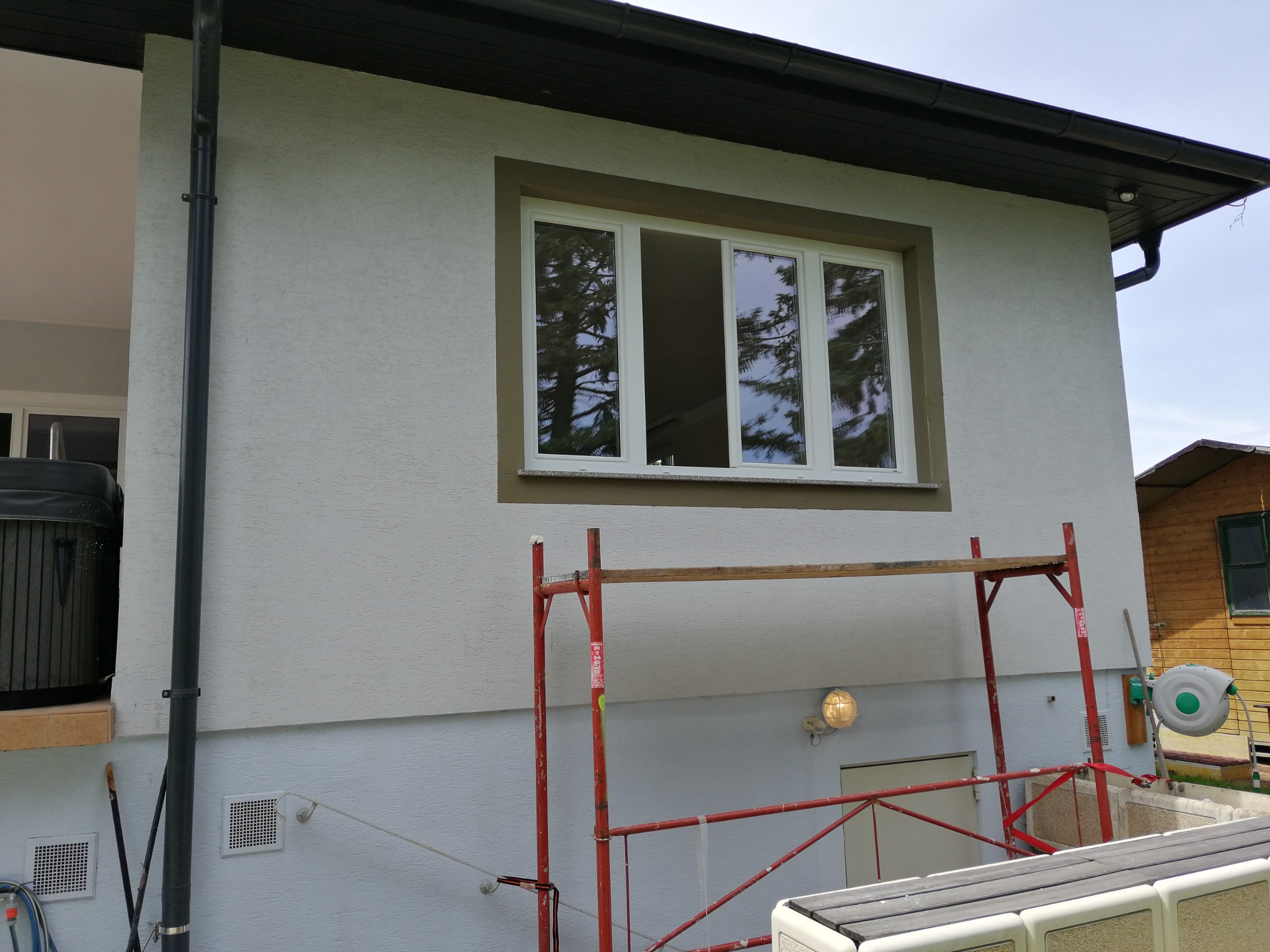 Dom w trakcie prac elewacyjnych: widoczne rusztowanie, okno z widocznym odbiciem drzew, fragment ocieplonej ściany z szarą strukturą tynku.