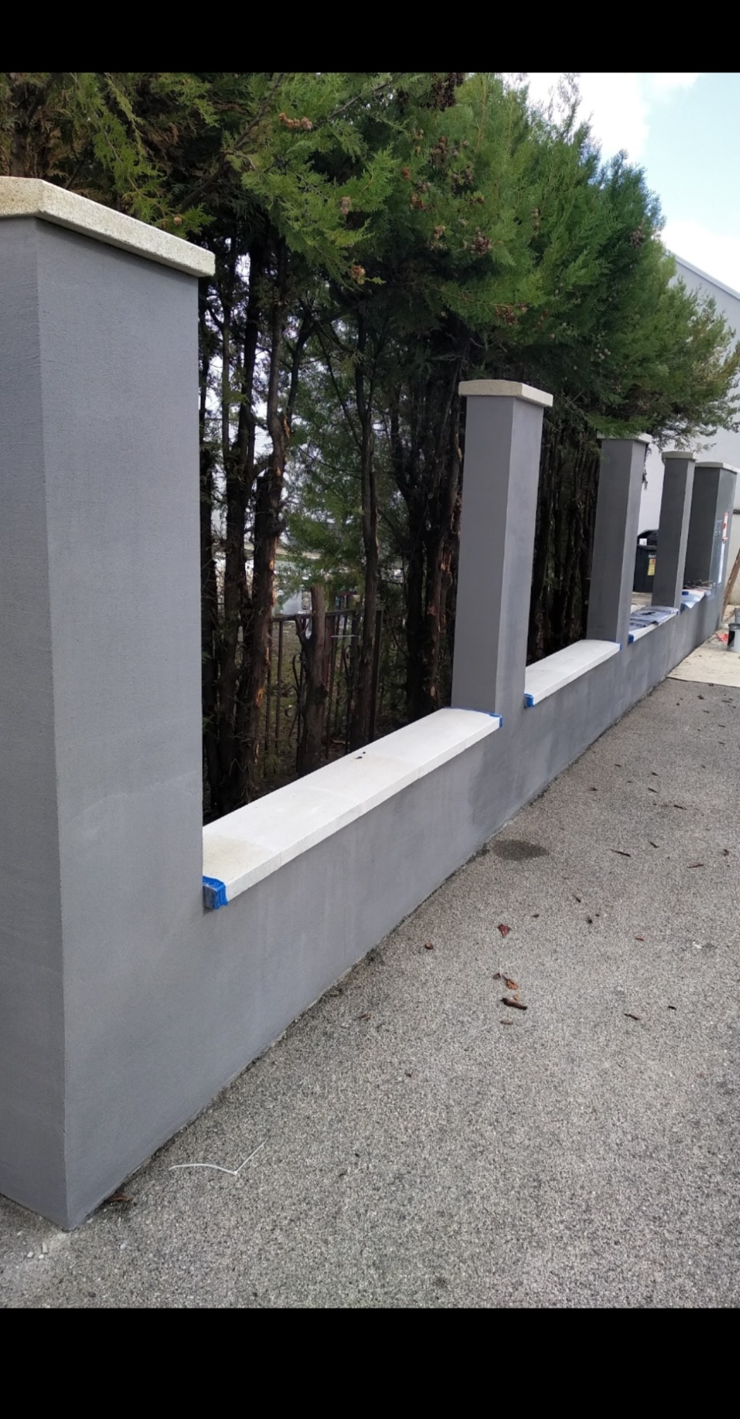 Nowoczesny, szary mur ogrodzeniowy z białymi elementami wykończeniowymi, widok z zewnątrz na tle zielonych drzew.