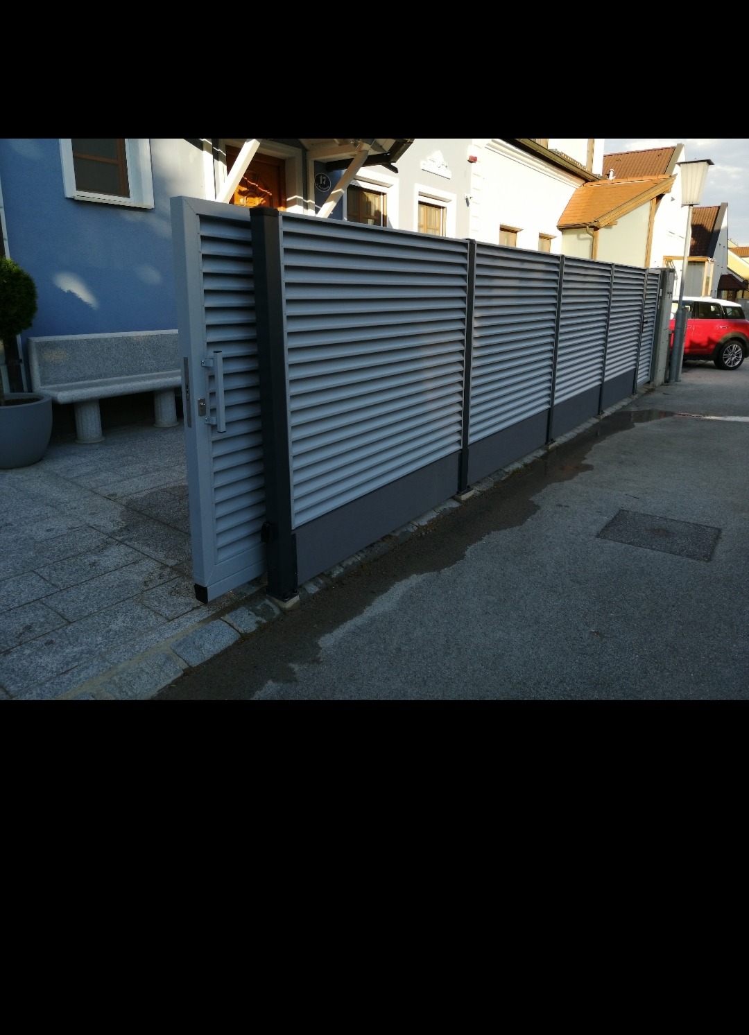 Nowoczesne, szare, przesuwne ogrodzenie lamelowe z widoczną częścią automatyki, na tle chodnika z kostki i ulicy, z domami w tle.