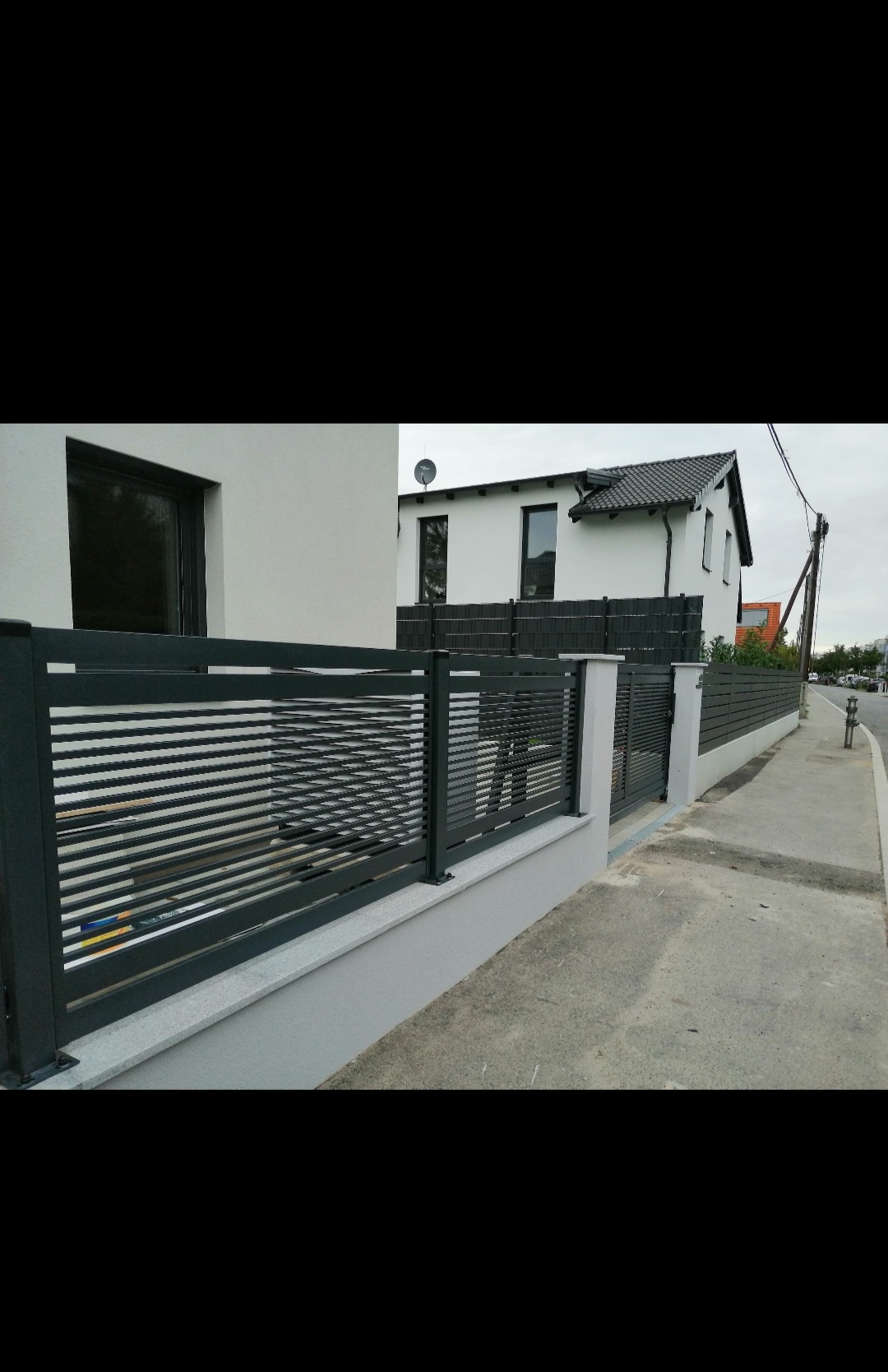 Nowoczesne ogrodzenie panelowe w kolorze antracytowym z poziomymi szczeblami, zamontowane na niskim, jasnoszarym murku, na tle białego domu z ciemną dachówką i szarą elewacją.