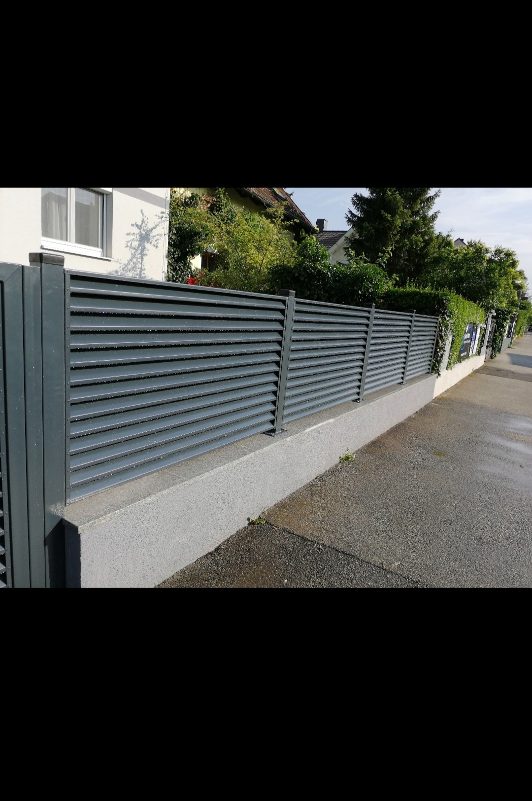 Nowoczesne, poziome, szare panele ogrodzeniowe zamontowane na niskiej, betonowej podmurówce, widoczne z perspektywy chodnika, z domem i zielenią w tle.