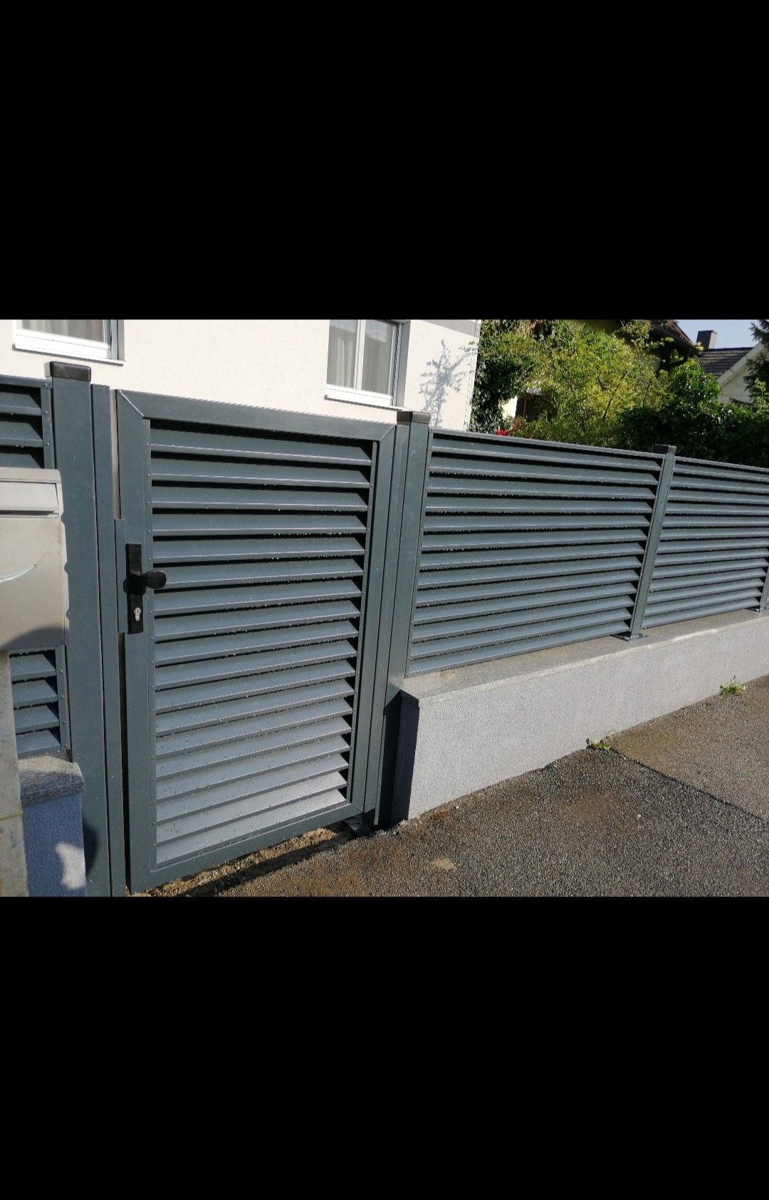 Nowoczesne, szare ogrodzenie lamelowe z furtką i słupkiem na betonowej podmurówce, widoczne z zewnątrz posesji, z fragmentem domu w tle.
